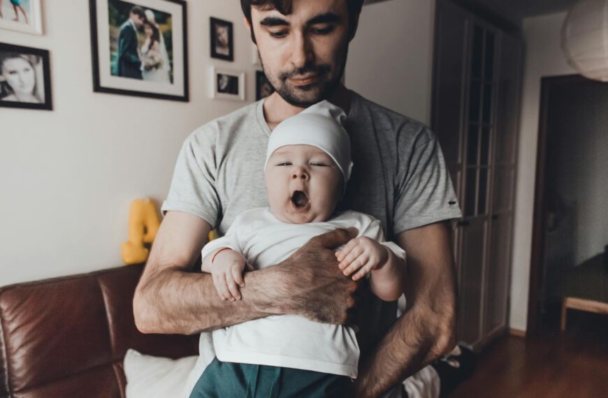 A father lovingly holds his yawning baby inside a warmly lit home, highlighting father-child bonding.