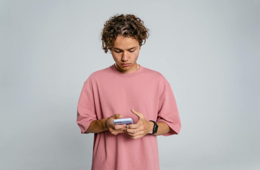 Teenage boy in pink shirt focuses on smartphone against gray background.