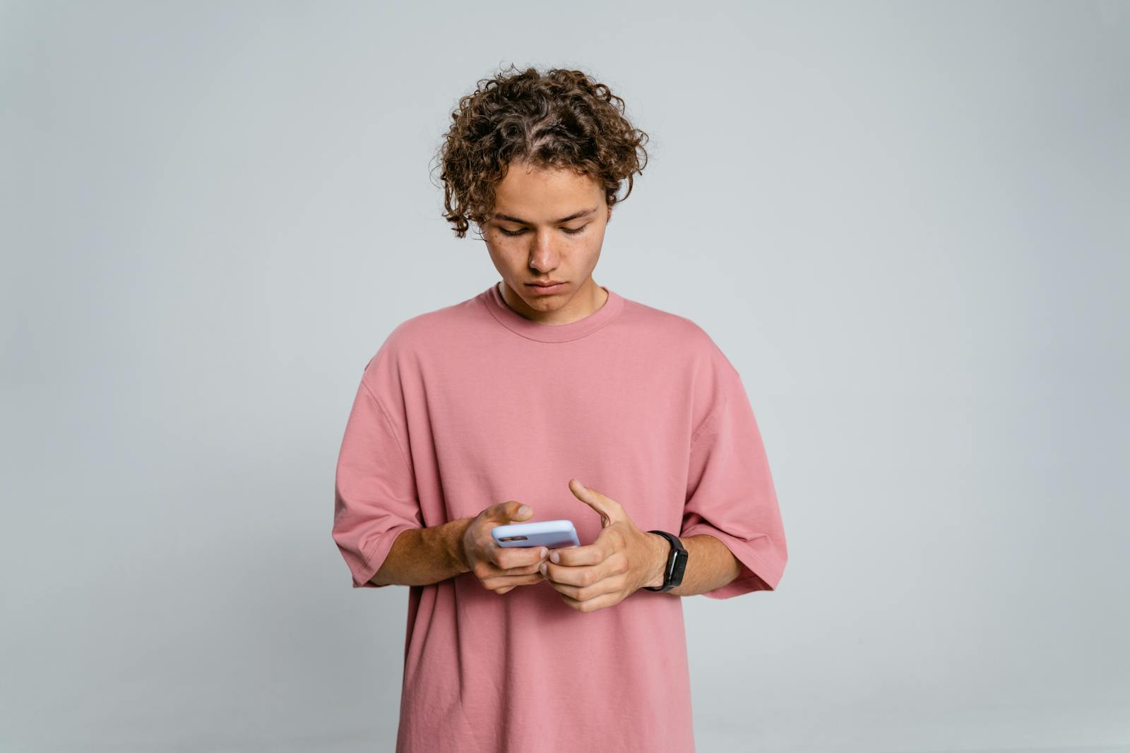 Teenage boy in pink shirt focuses on smartphone against gray background.