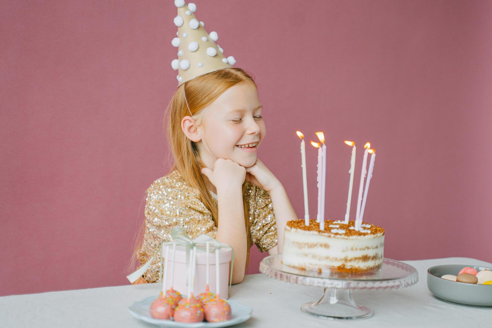 A young girl in a party hat smiles at her birthday cake with candles lit, ready to make a wish.