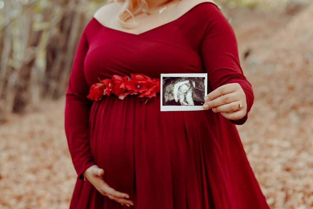 Expecting mother in red dress showing ultrasound in a serene autumn forest setting.