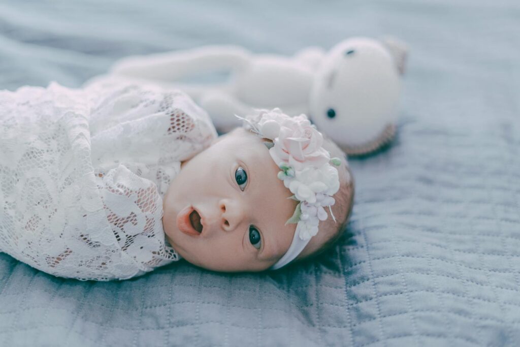 Charming newborn wearing a floral headband and swaddled in lace, resting peacefully with a plush toy beside.