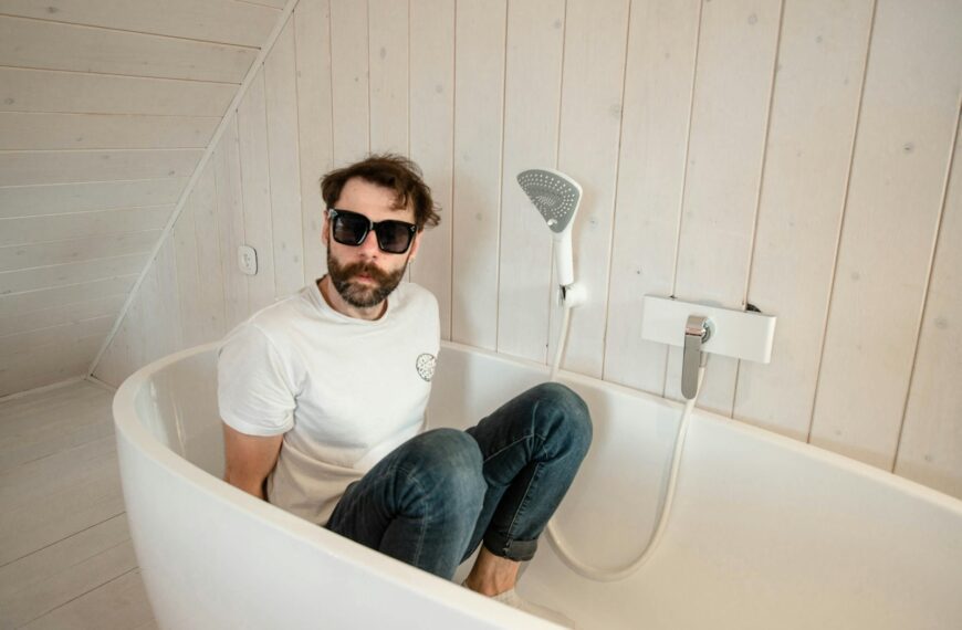 man in white crew neck t-shirt and blue denim jeans sitting on white ceramic bathtub