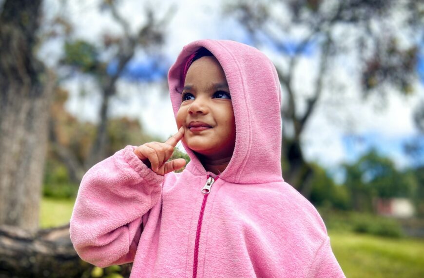 a little girl in a pink jacket standing in front of a tree