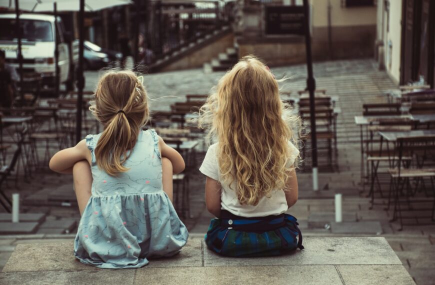 two girls sitting on concrete pavement facing on dining tables during daytime