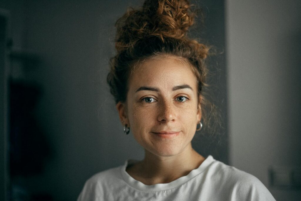 Portrait of a smiling woman with curly hair and freckles in soft light, wearing a white t-shirt indoors.