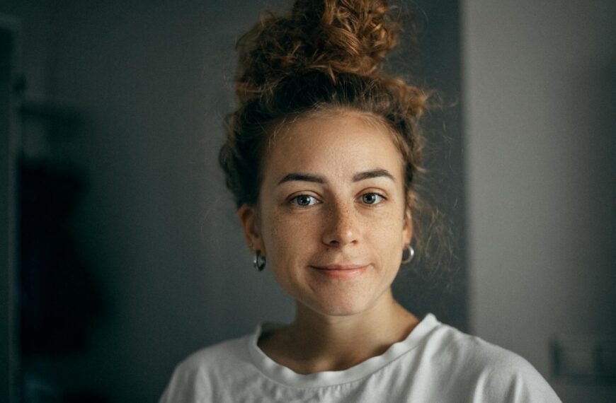 Portrait of a smiling woman with curly hair and freckles in soft light, wearing a white t-shirt indoors.