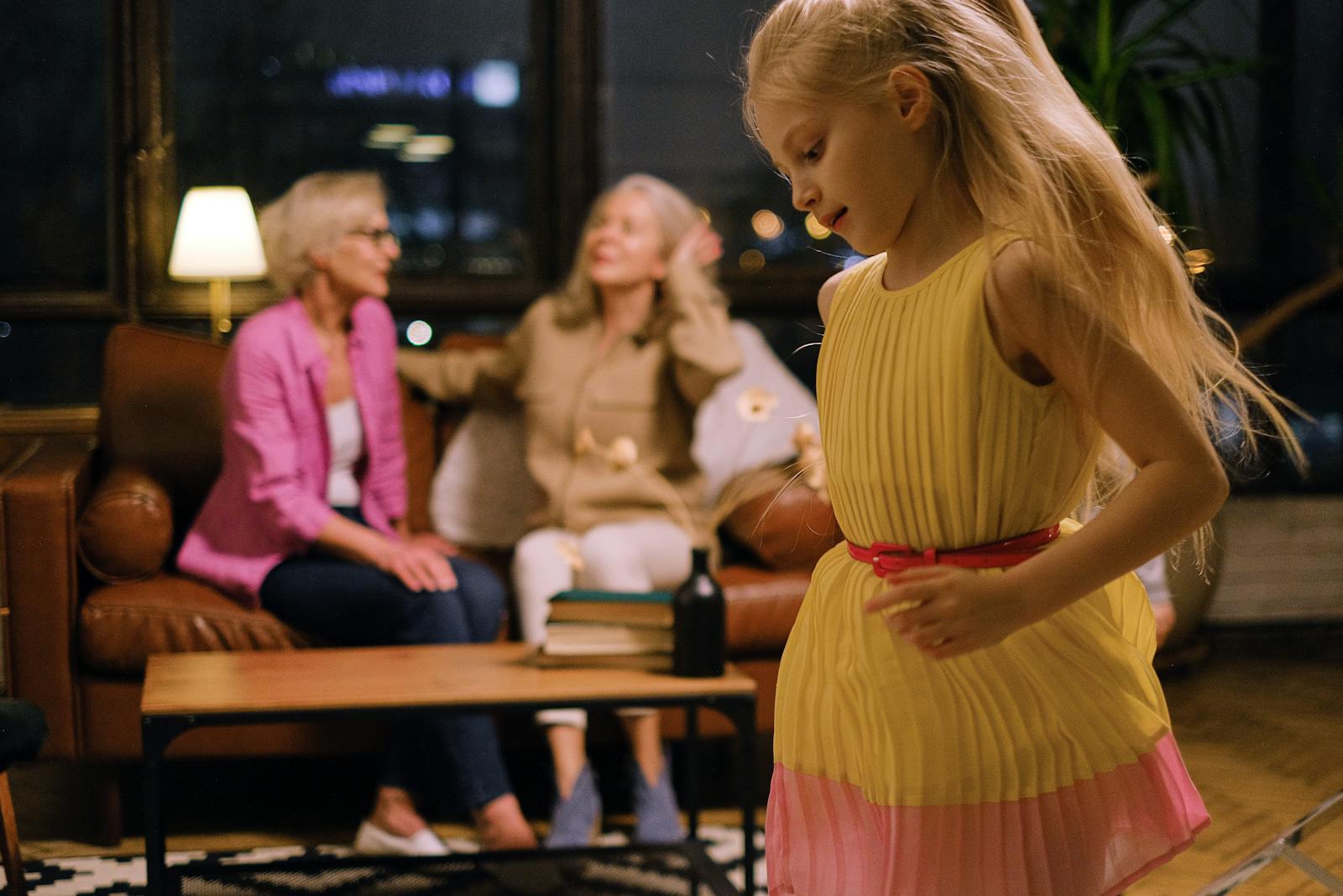 A joyful young girl in a yellow dress dances inside while family members relax on the couch.