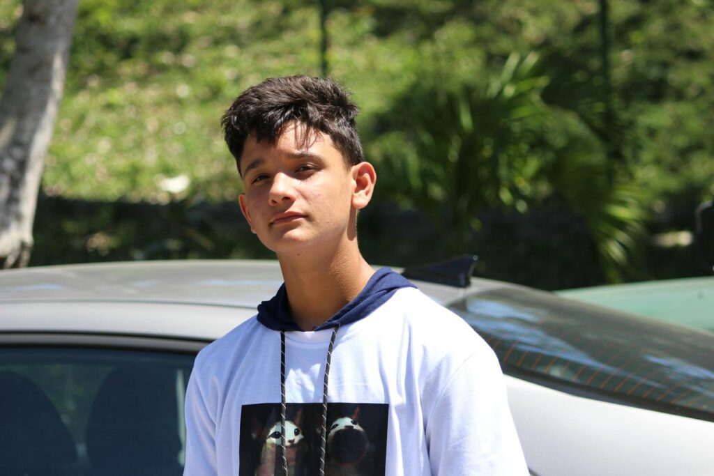 Teenage boy standing by a car outdoors on a sunny day with a thoughtful expression.