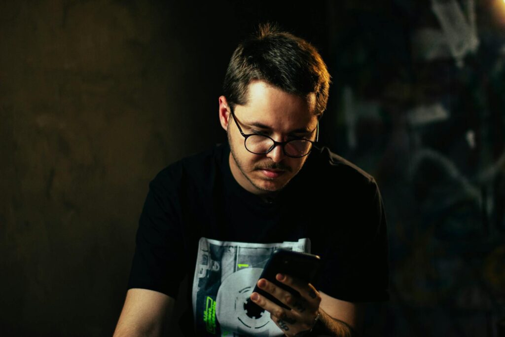 Young man with eyeglasses focused on smartphone indoors in moody lighting.