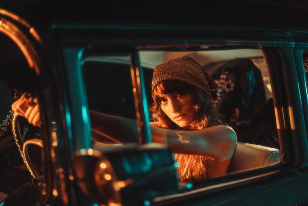 A young woman in a beanie poses confidently in a vintage car, illuminated by cinematic teal and orange lighting.