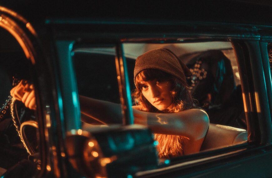 A young woman in a beanie poses confidently in a vintage car, illuminated by cinematic teal and orange lighting.