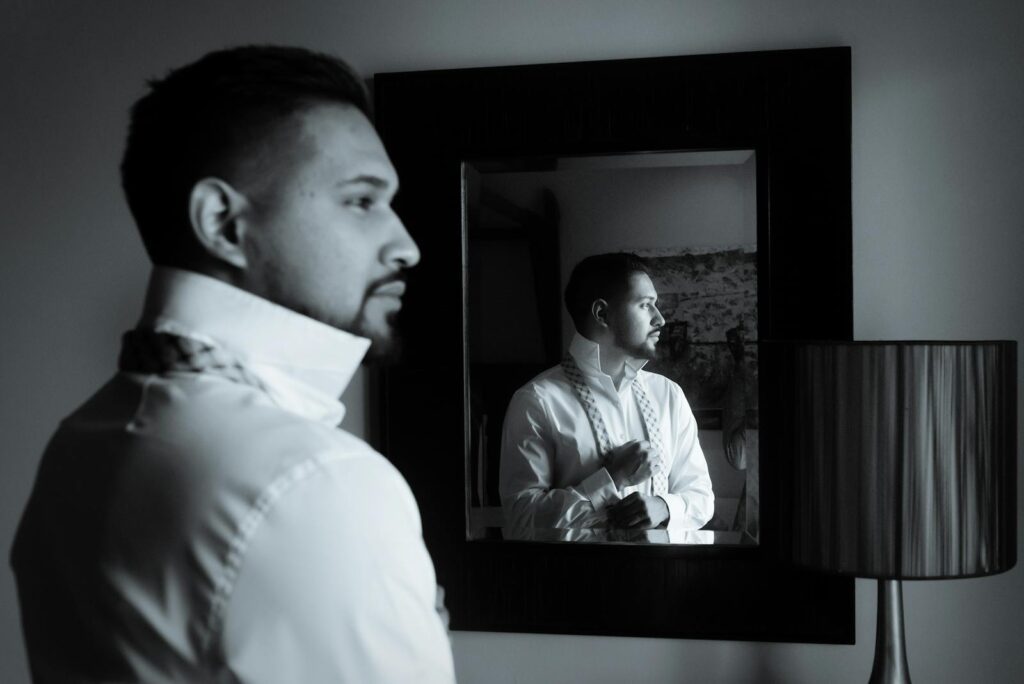 A black and white image of a man adjusting his bow tie while looking into a mirror.
