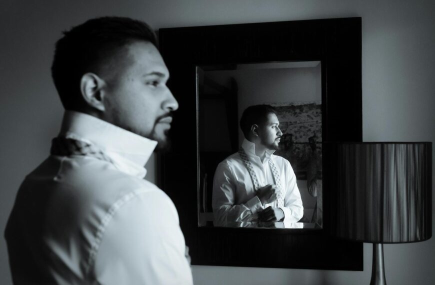 A black and white image of a man adjusting his bow tie while looking into a mirror.