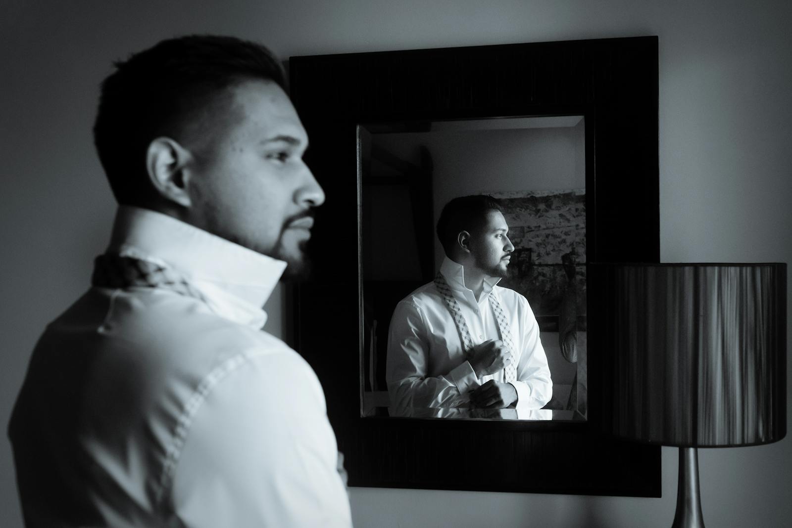 A black and white image of a man adjusting his bow tie while looking into a mirror.