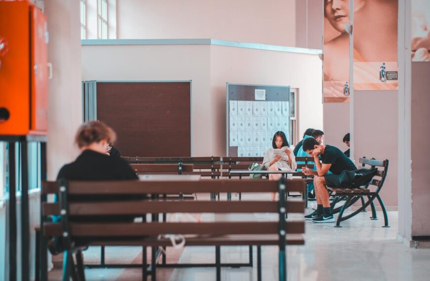 People sitting on benches in a school lobby, waiting and using smartphones, in a quiet indoor setting.