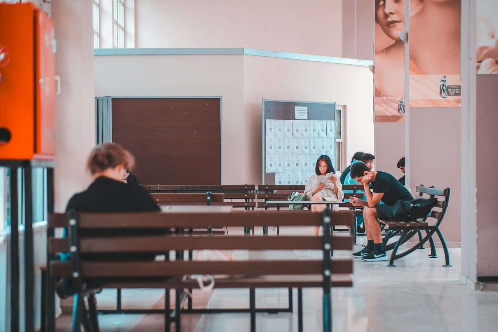 People sitting on benches in a school lobby, waiting and using smartphones, in a quiet indoor setting.