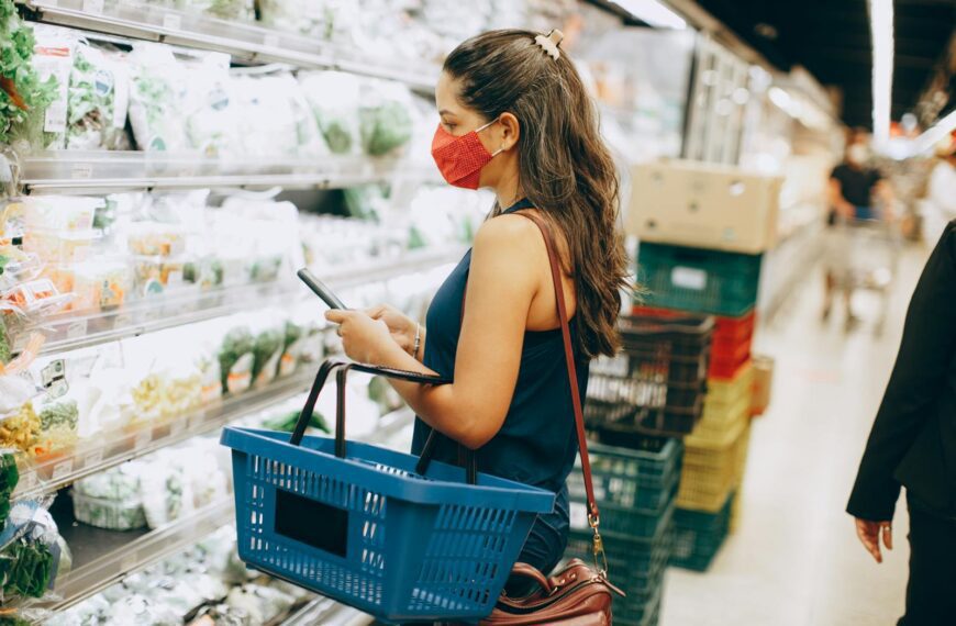 A woman wearing a mask shops for fresh produce while using a smartphone in a grocery store.