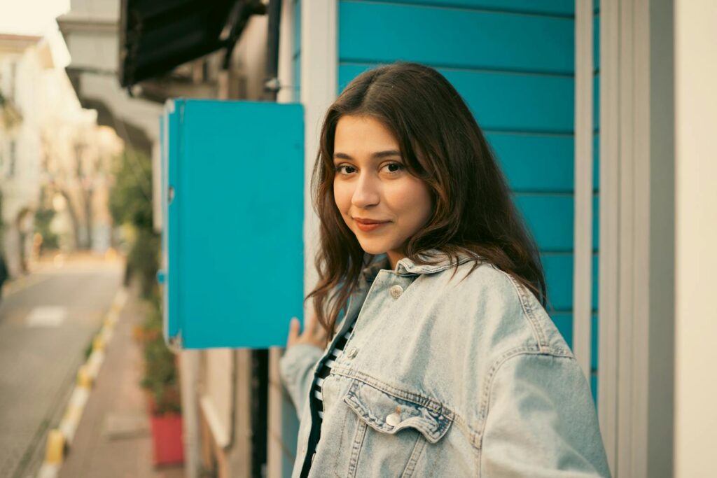 Portrait of a young woman in a denim jacket outdoors by a blue wall.