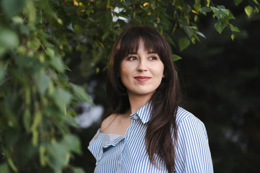 Portrait of a young woman in a striped shirt standing outdoors, surrounded by nature.
