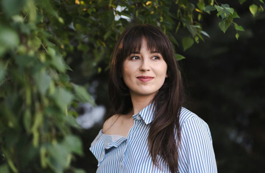 Portrait of a young woman in a striped shirt standing outdoors, surrounded by nature.