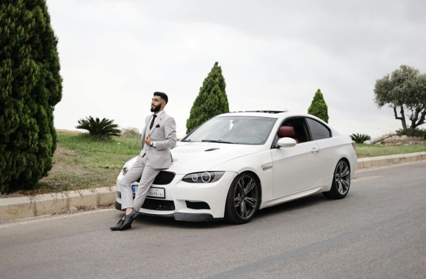Elegant man in a suit leans on a white sports car, set against a scenic outdoor backdrop.