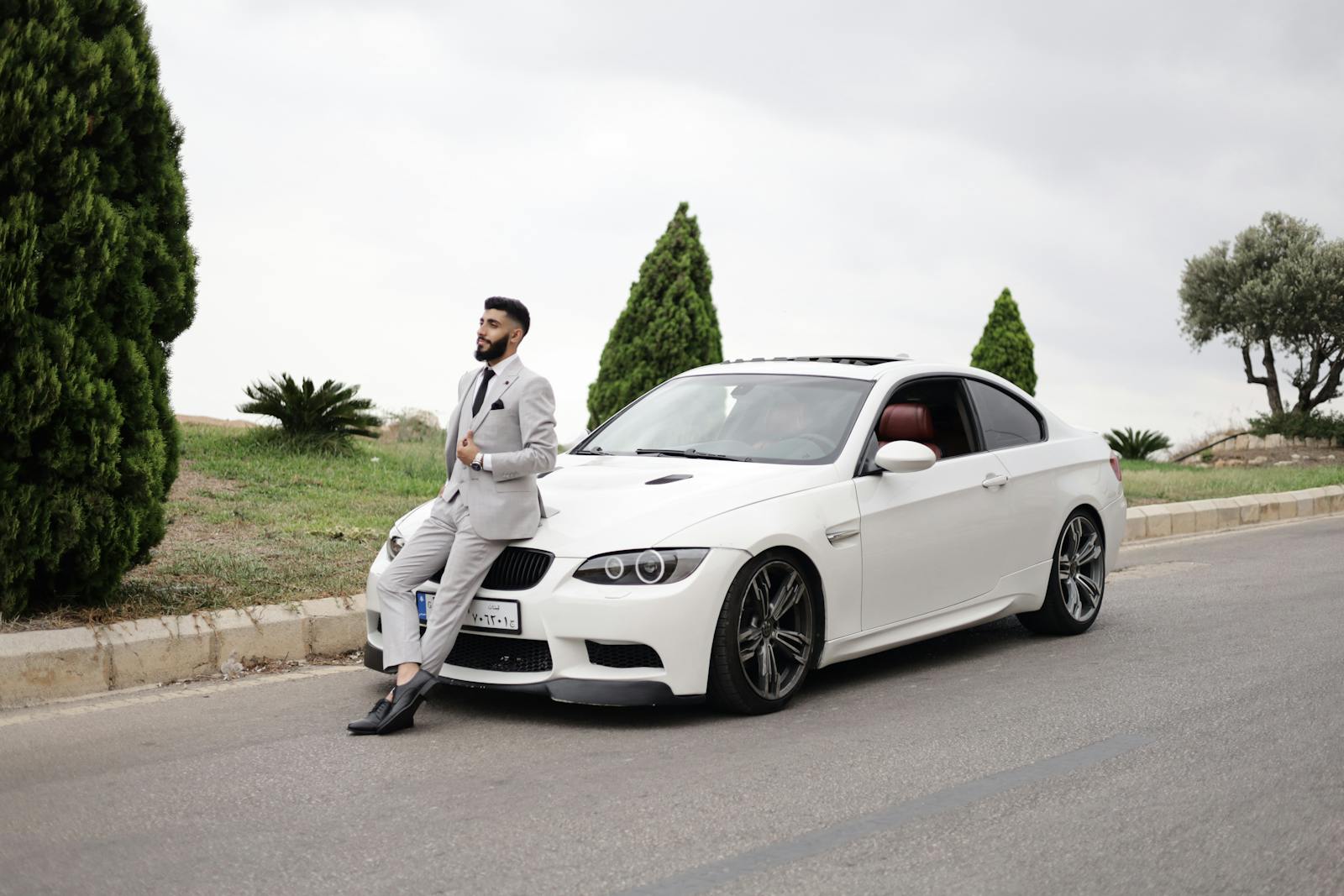 Elegant man in a suit leans on a white sports car, set against a scenic outdoor backdrop.