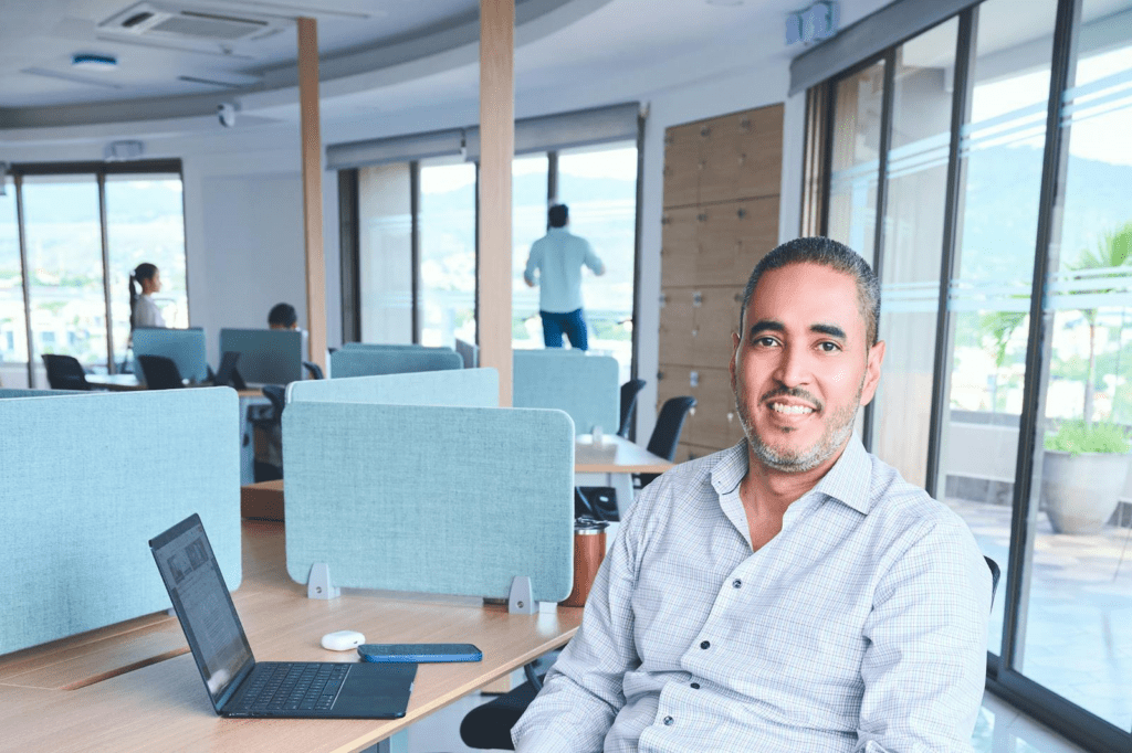 A smiling man sits in a bright, modern co-working space with a laptop.