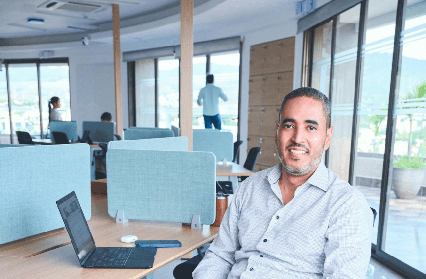 A smiling man sits in a bright, modern co-working space with a laptop.