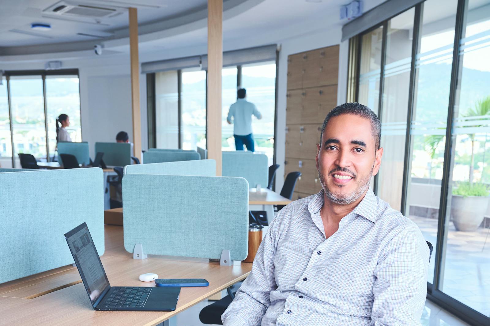 A smiling man sits in a bright, modern co-working space with a laptop.