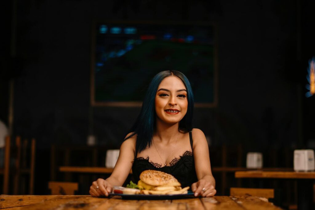 A young woman with blue hair and braces smiles while holding a burger in a cozy restaurant setting.