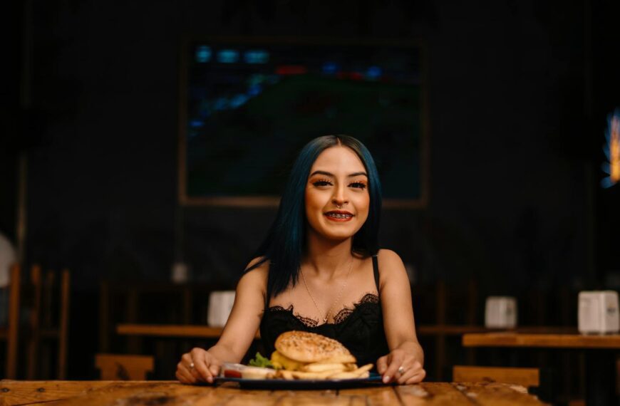 A young woman with blue hair and braces smiles while holding a burger in a cozy restaurant setting.