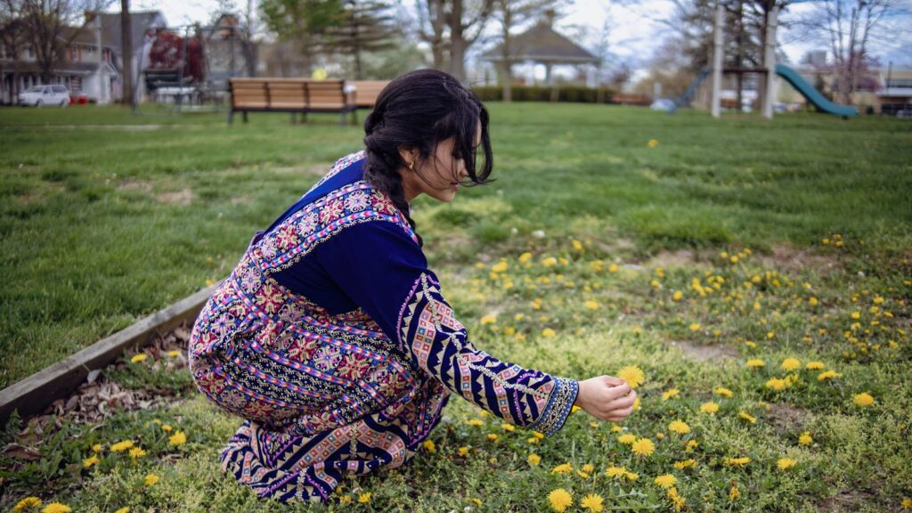 Woman in a vibrant dress picking dandelions on a sunny day in a park.