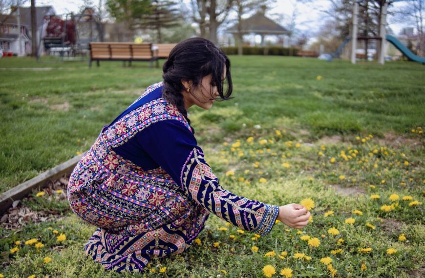 Woman in a vibrant dress picking dandelions on a sunny day in a park.