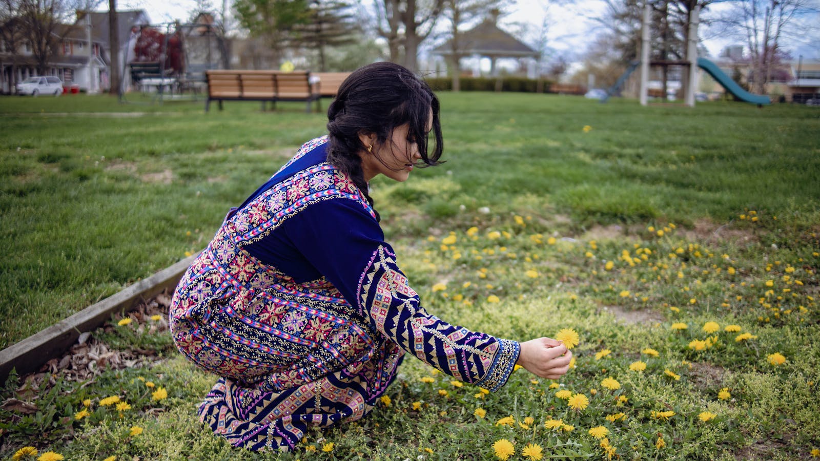 Woman in a vibrant dress picking dandelions on a sunny day in a park.