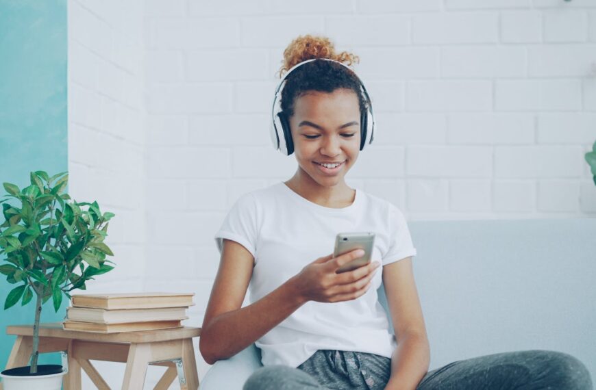 Smiling teen girl enjoying music on headphones while texting on smartphone at home.