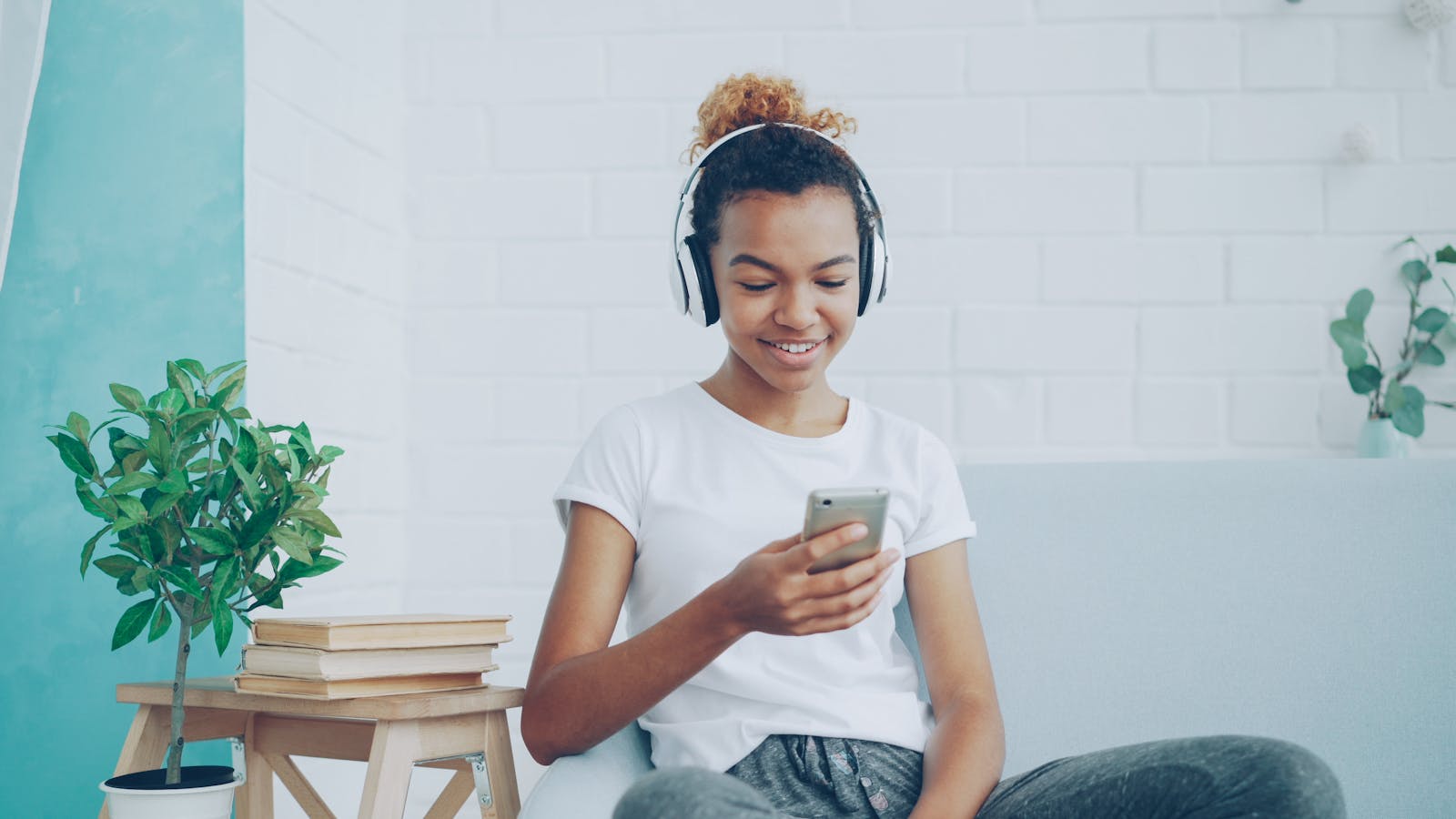 Smiling teen girl enjoying music on headphones while texting on smartphone at home.