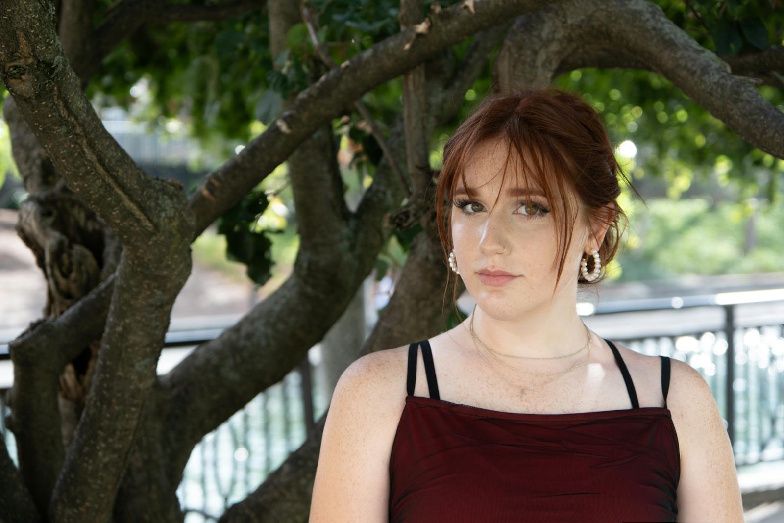 Beautiful young woman with red hair posing outdoors in front of a tree on a sunny spring day.