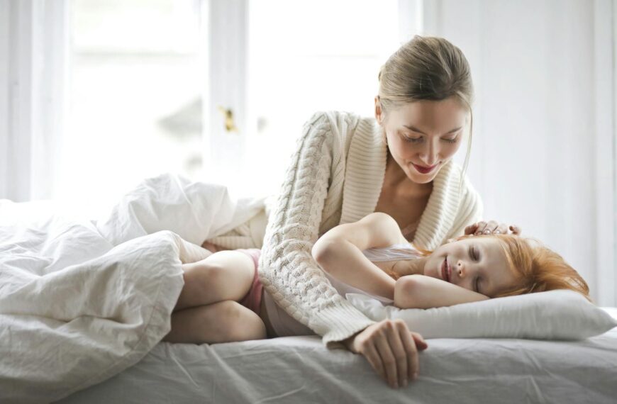 Cheerful mother caressing child while girl sleeping on bed in cozy room at home