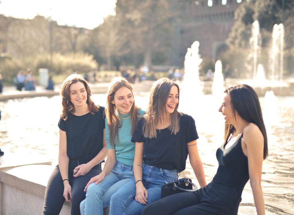 Four friends laughing and chatting outdoors by a water fountain on a sunny day.