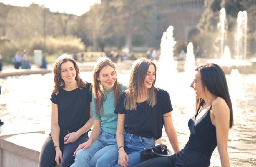 Four friends laughing and chatting outdoors by a water fountain on a sunny day.