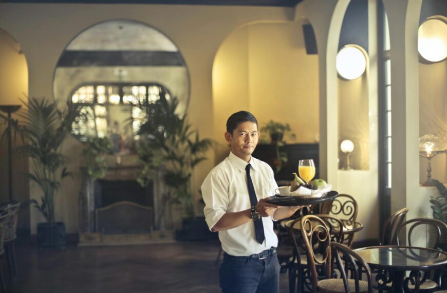 Confident young ethnic waiter in elegant clothes holding tray with food and drinks and looking at camera while serving tables in stylish restaurant