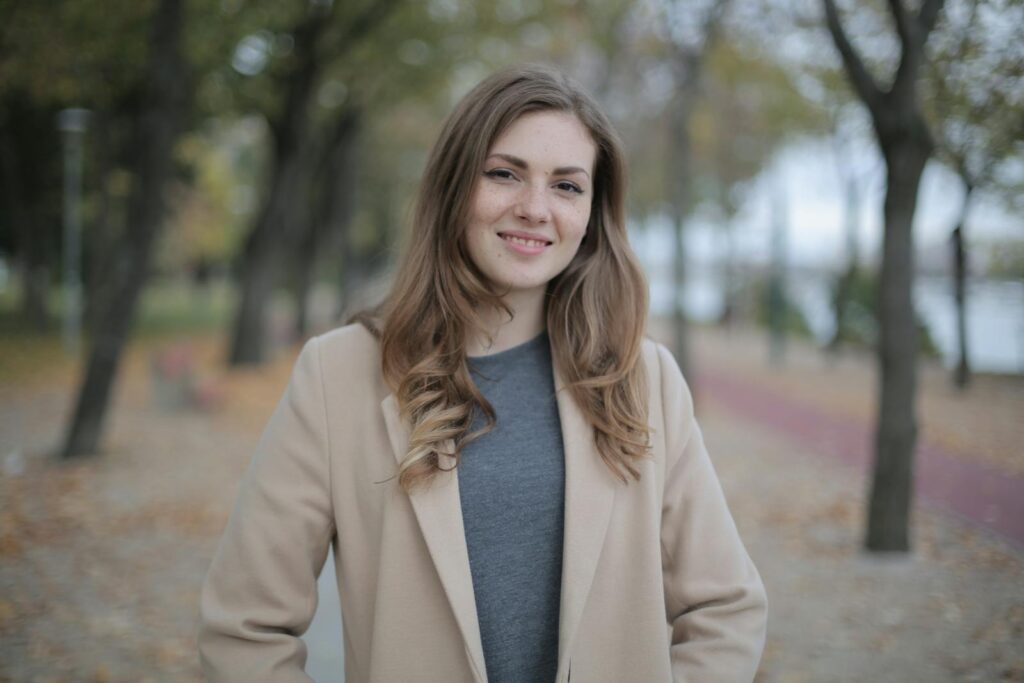 A young woman in a beige coat smiles warmly while standing outdoors during fall.
