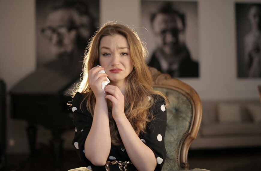 A woman sitting in a vintage chair indoors, showing emotions and holding tissues.