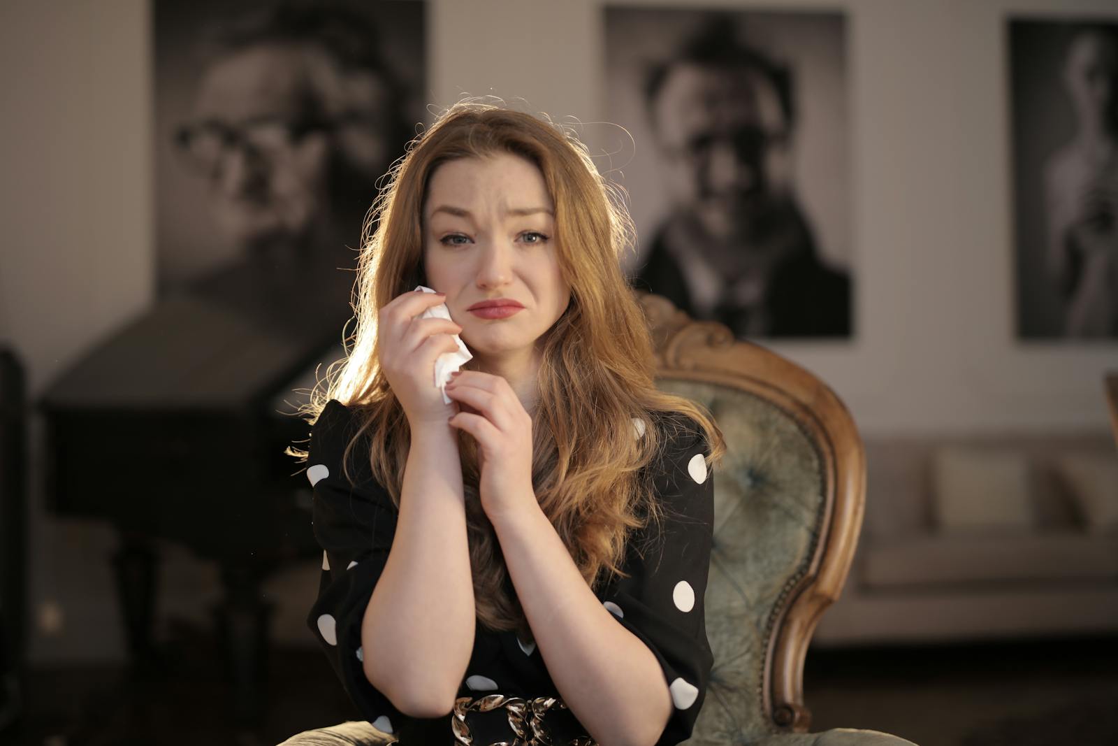A woman sitting in a vintage chair indoors, showing emotions and holding tissues.