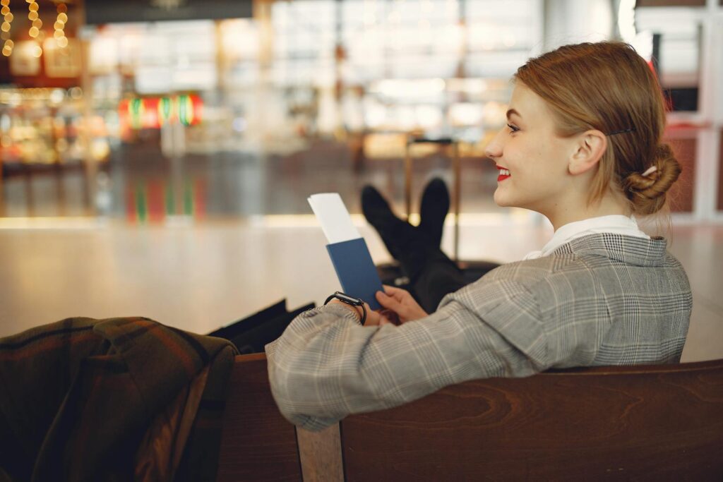 Side view of cheerful female student in checkered jacket smiling away while chilling in hall with outstretched legs and passport in hands