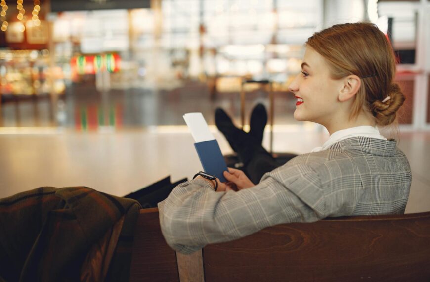 Side view of cheerful female student in checkered jacket smiling away while chilling in hall with outstretched legs and passport in hands