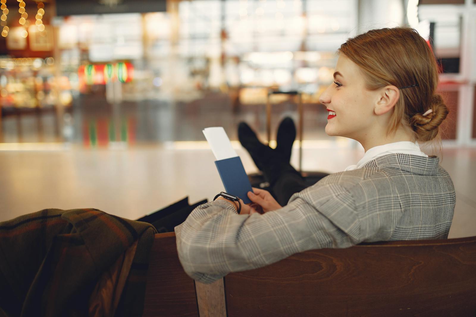 Side view of cheerful female student in checkered jacket smiling away while chilling in hall with outstretched legs and passport in hands