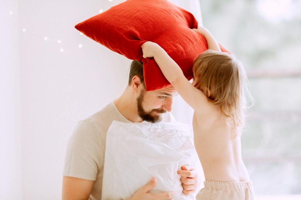 A joyous moment of a father and daughter engaging in a playful pillow fight indoors.
