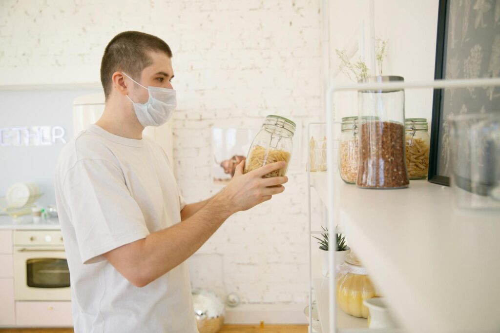A young man with a facemask in a modern kitchen reviewing pantry items.